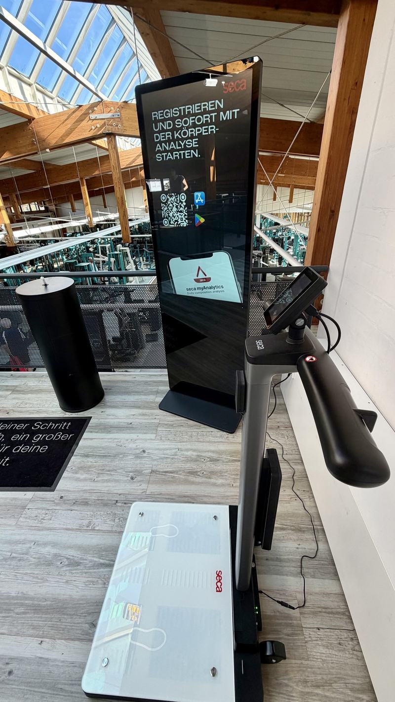 seca TRU auf der Galerie: Waage mit Handgriffen und Companion-Stele, im Hintergrund die Geräte der Trainingsfläche